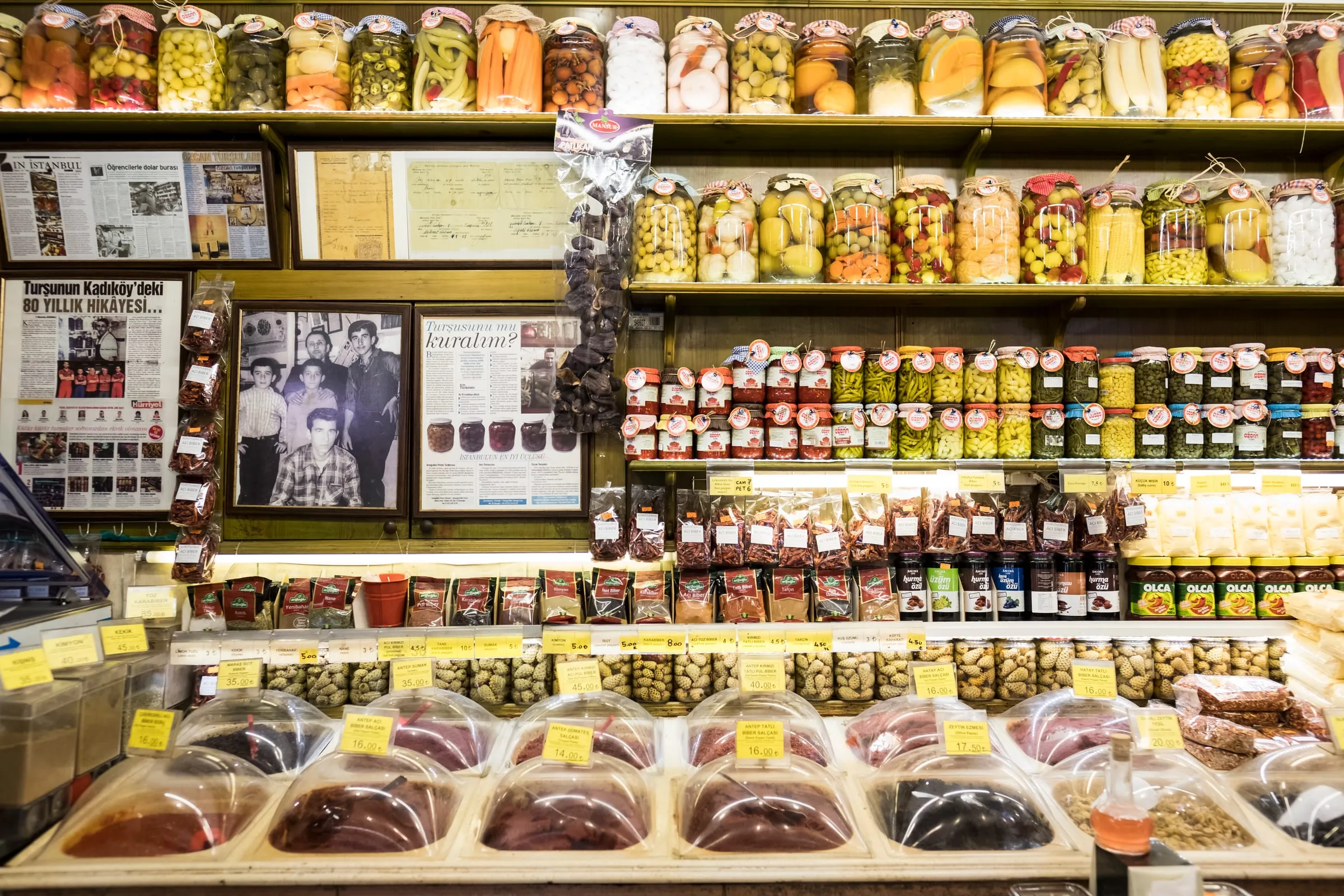 Traditional shop counter with labeled bins of Maraş pepper, isot, sumac, and pepper pastes under shelves of pickle jars