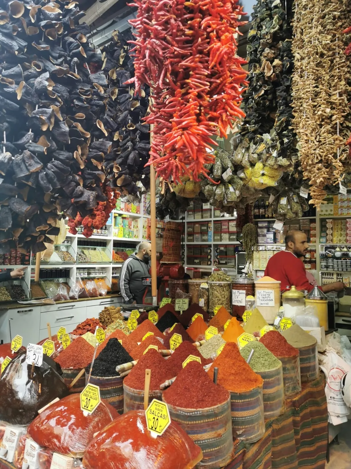 Aktar shop with strings of dried peppers and eggplants hanging over mounds of pepper paste and spices with handwritten price tags
