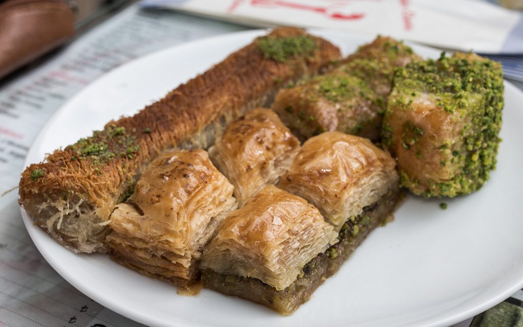 An assortment of Turkish baklava on a white plate, topped with ground pistachio, including a shredded kadayıf roll and layered fıstıklı squares