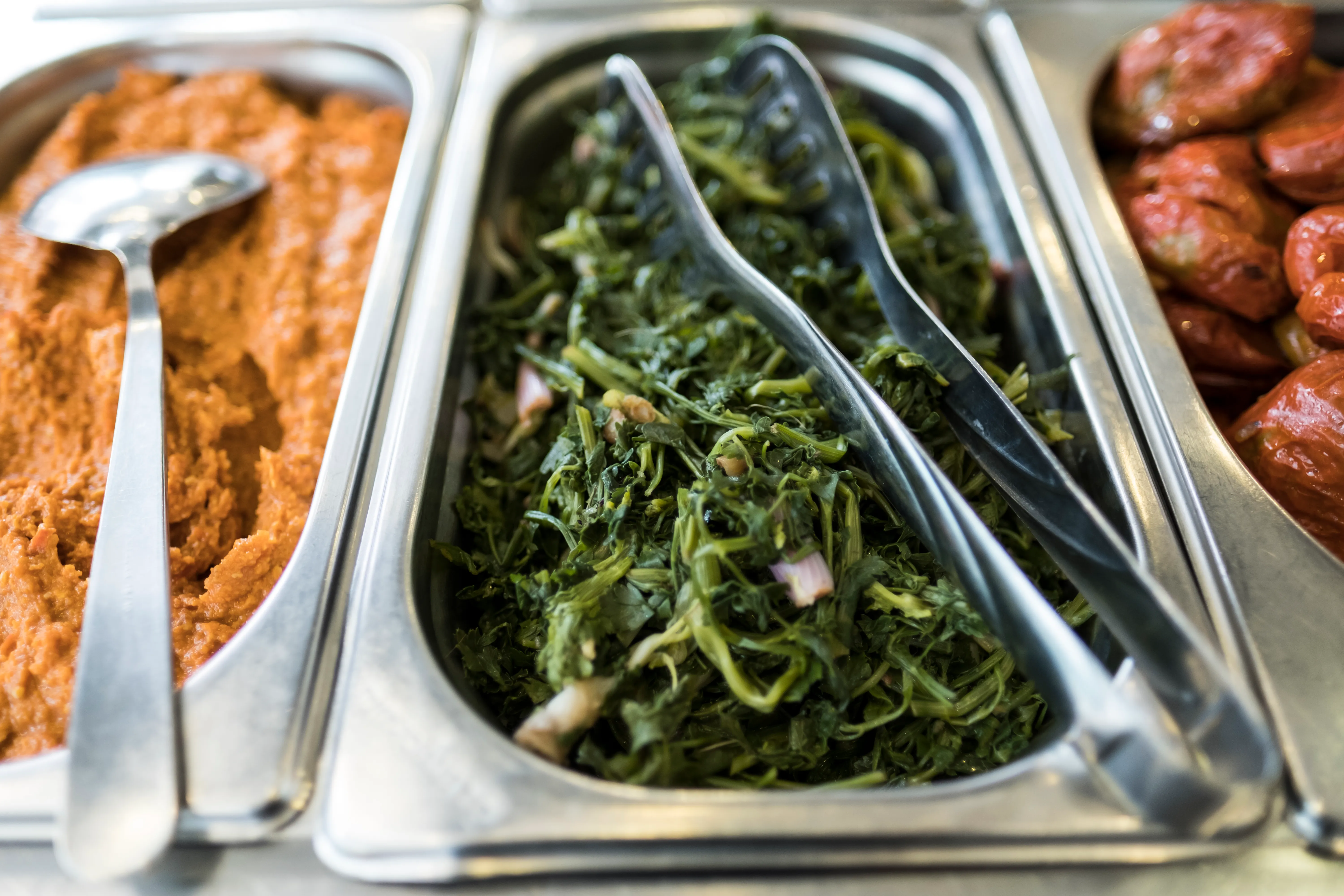 Cooked leafy greens and a serving spoon in a metal tray beside other dishes