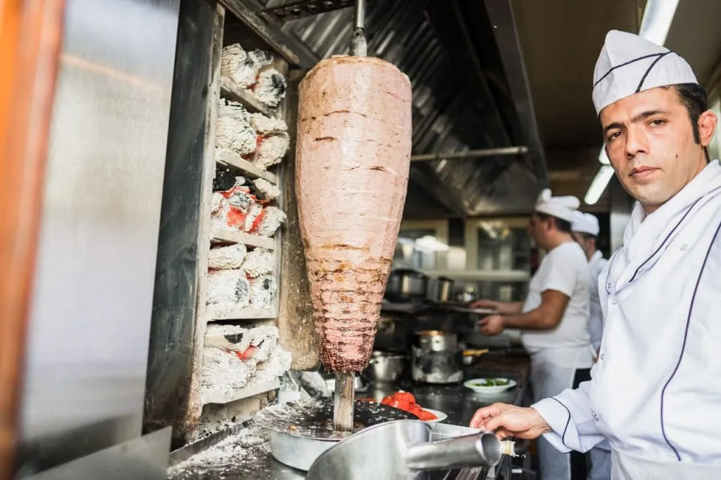 Chef beside large doner kebab meat on rotisserie being cooked by wood in istanbul restaurant kitchen