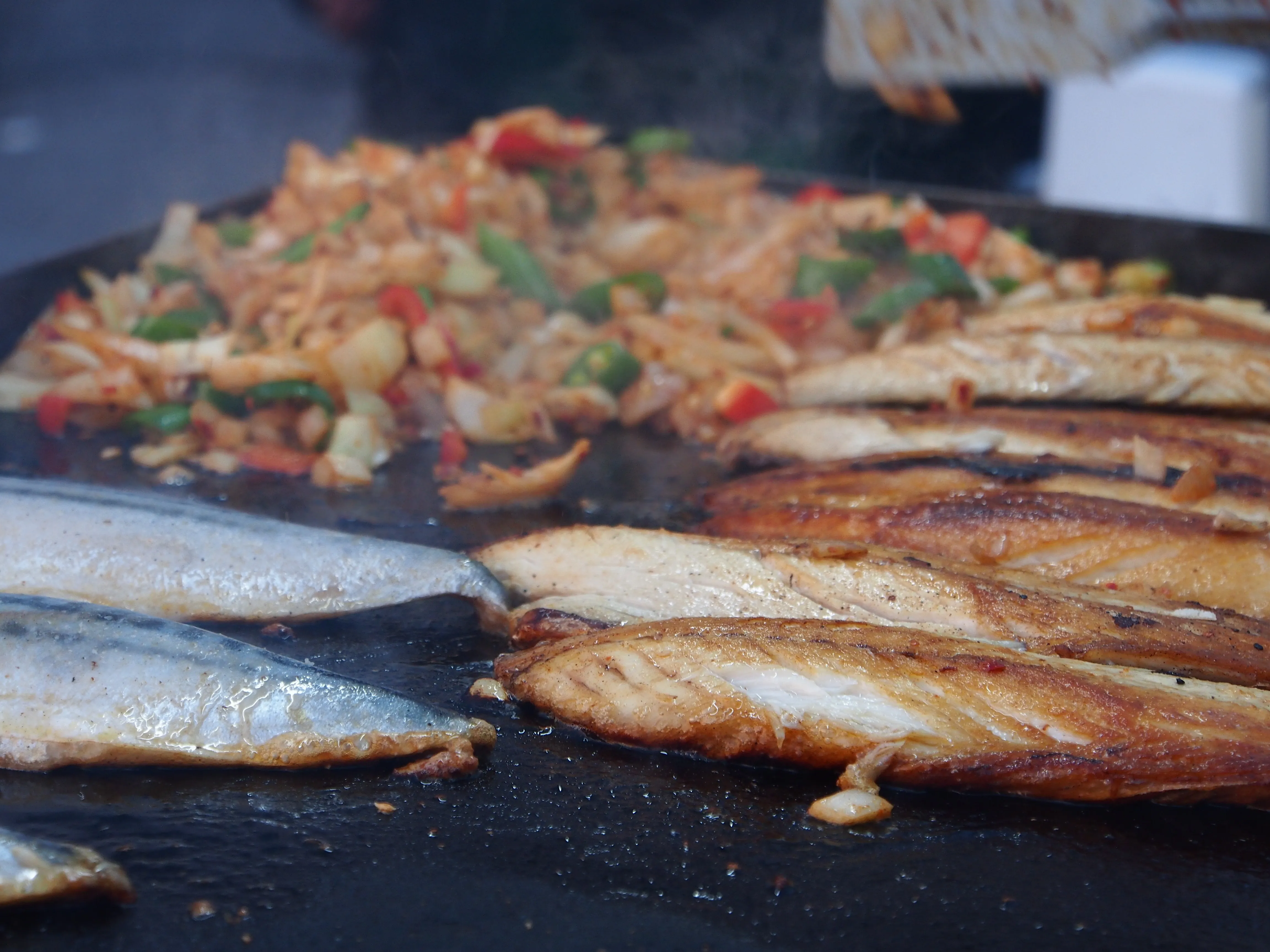 Fish fillets brown on a hot flat griddle beside chopped tomato and herbs