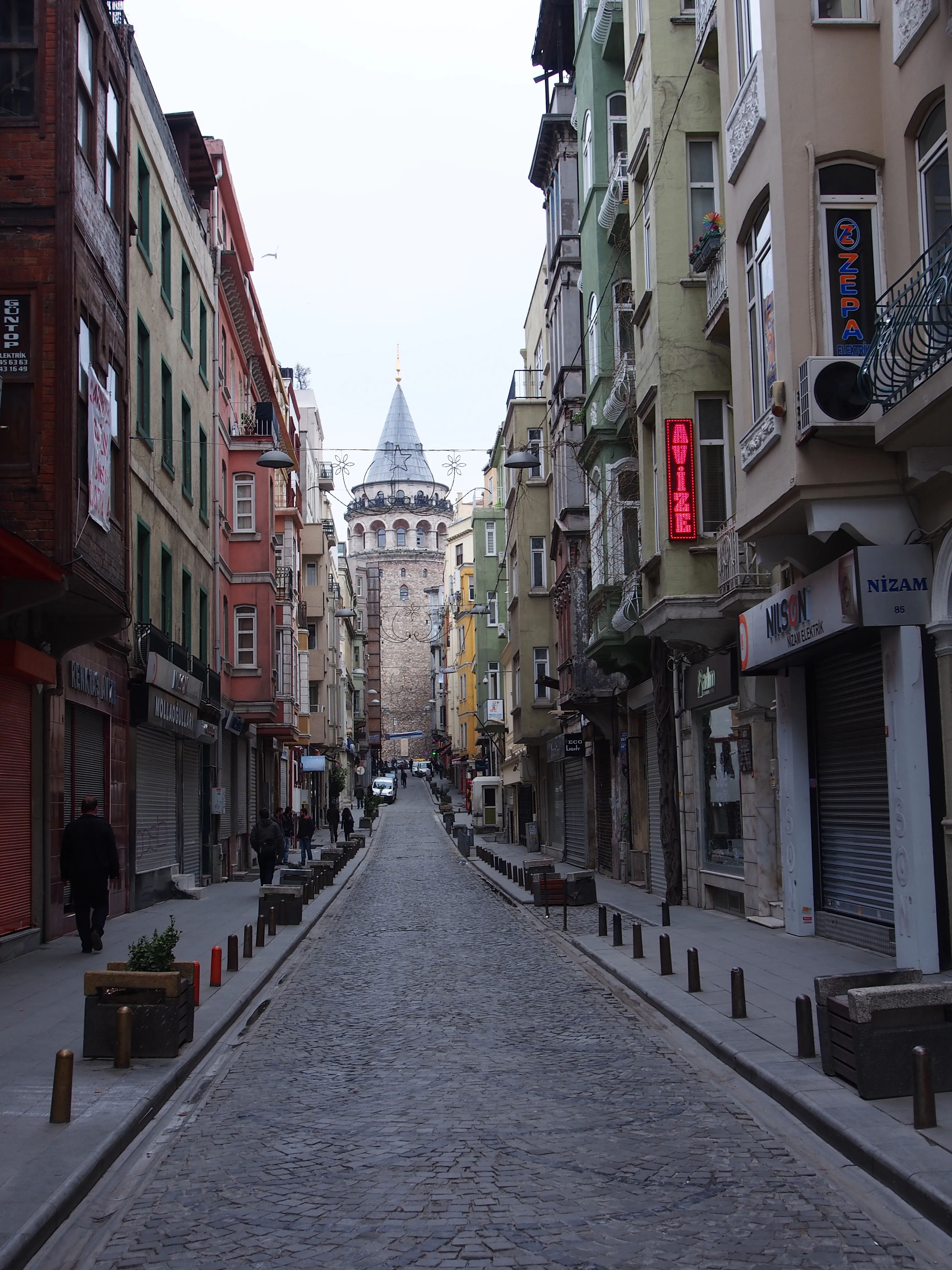A cobbled street climbs between buildings toward Galata Tower