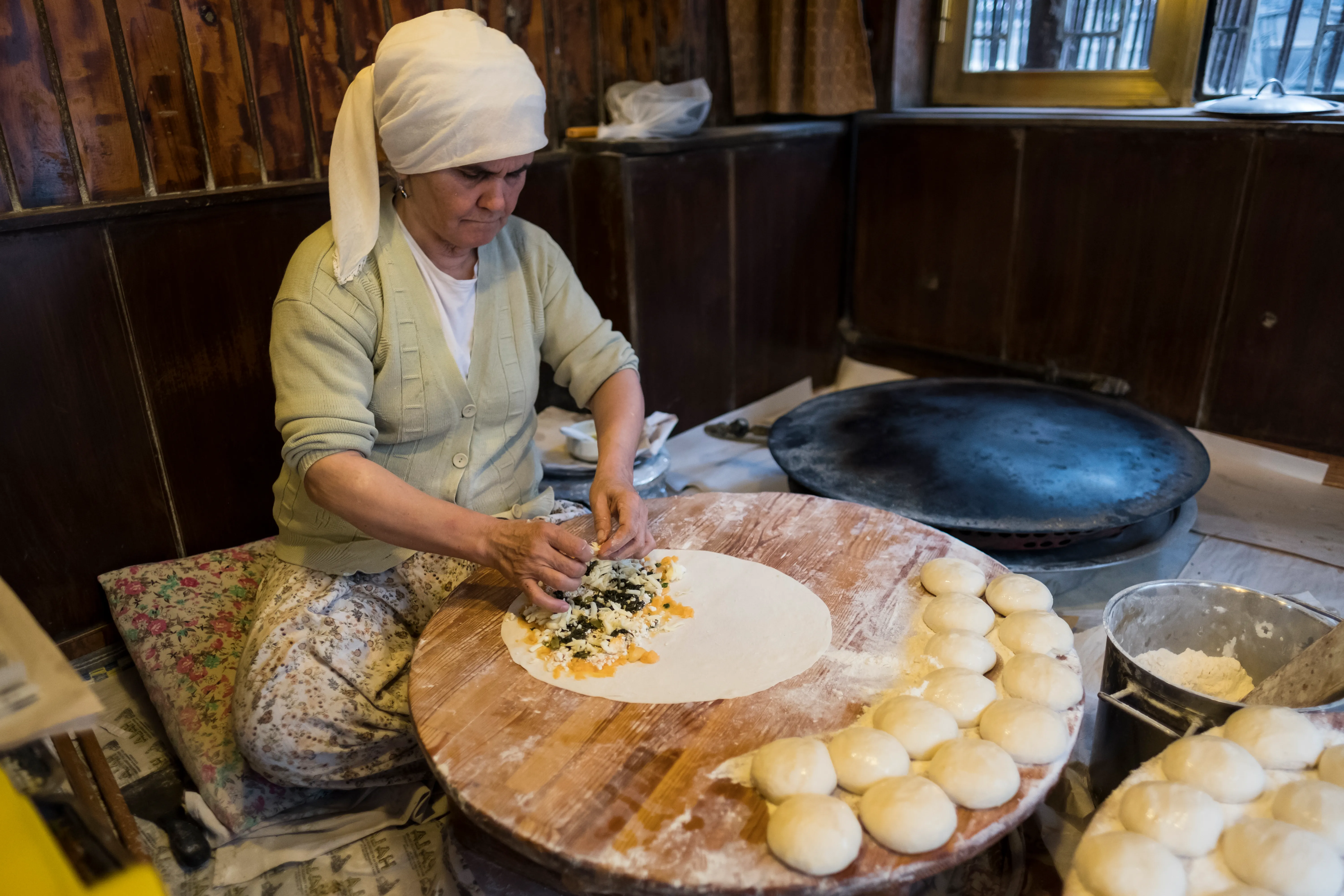 A cook adds filling to thin round dough at a low work surface beside a griddle