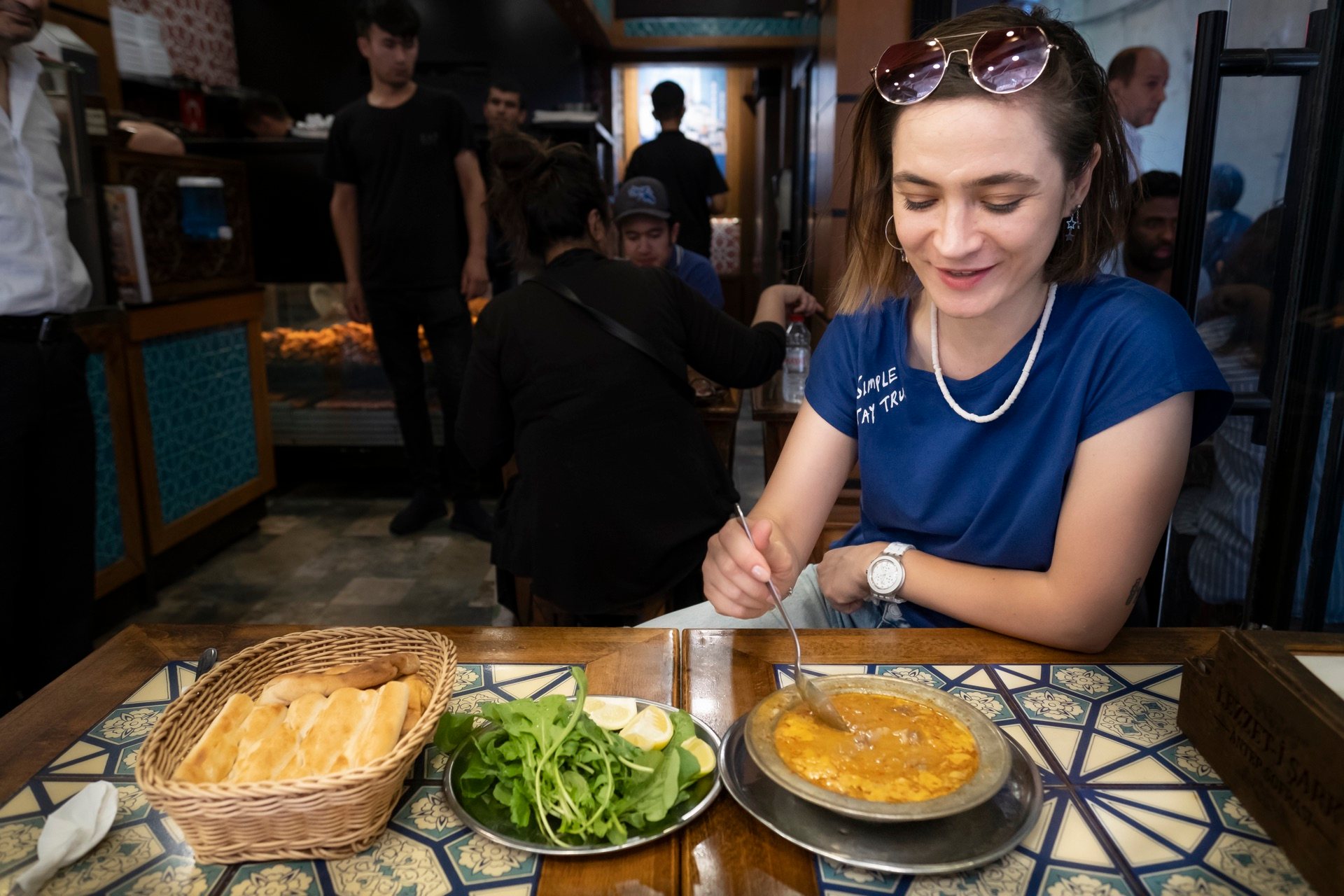 Smiling guest at a Turkish lokanta table with a metal plate, arugula salad, and a basket of bread