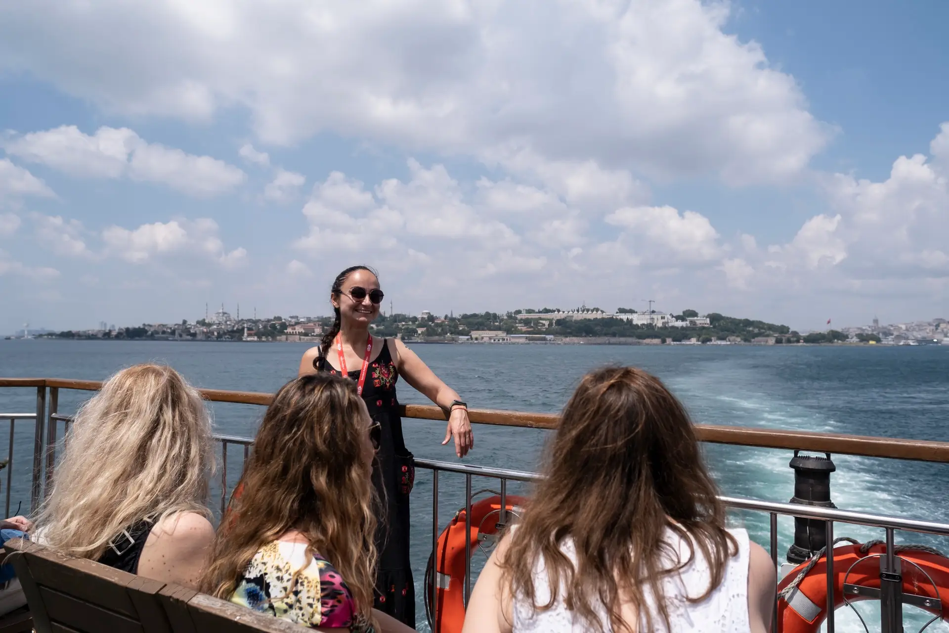 Tour guide on the ferry stern talking to seated guests, with Istanbul's historic peninsula and Topkapı Palace across the water