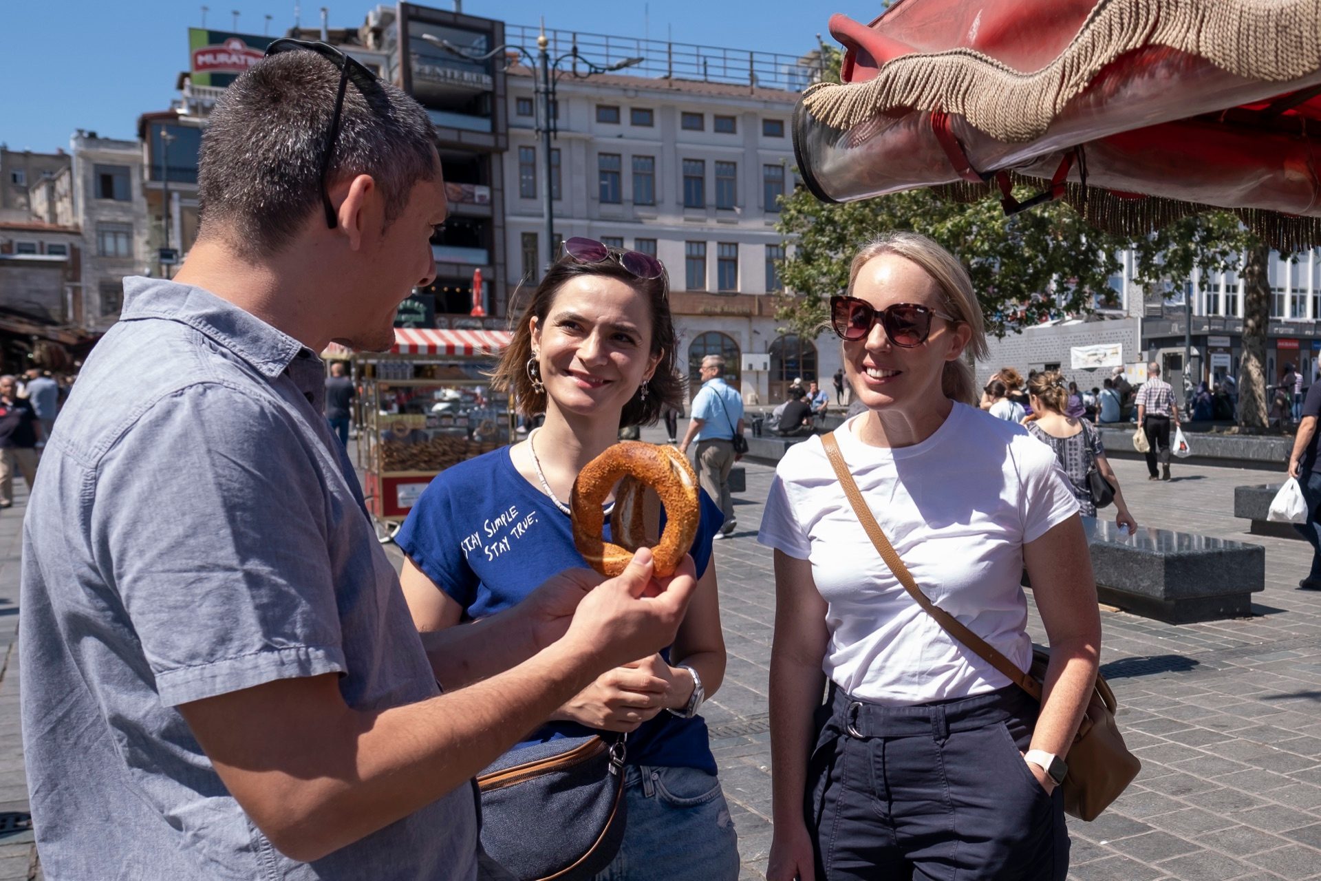 Tour guide holding a round Turkish simit explaining it to two guests in a sunny square
