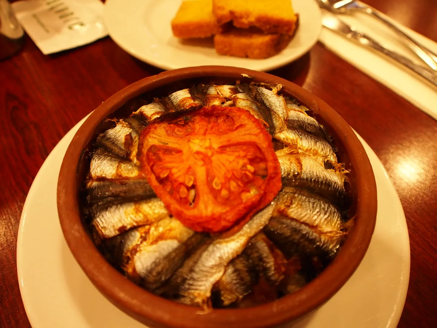 baked anchovies arranged in a ring around a tomato slice in a clay dish, with cornbread alongside