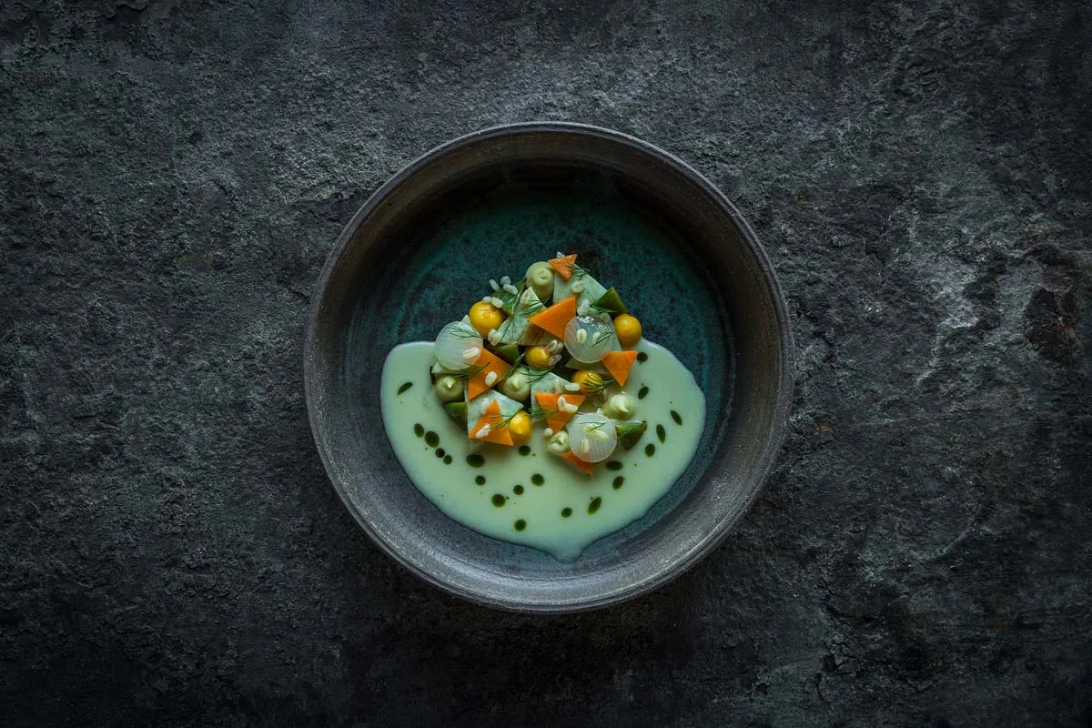Plated dish of carrot, corn, and pearl onions in pale green sauce on dark stone