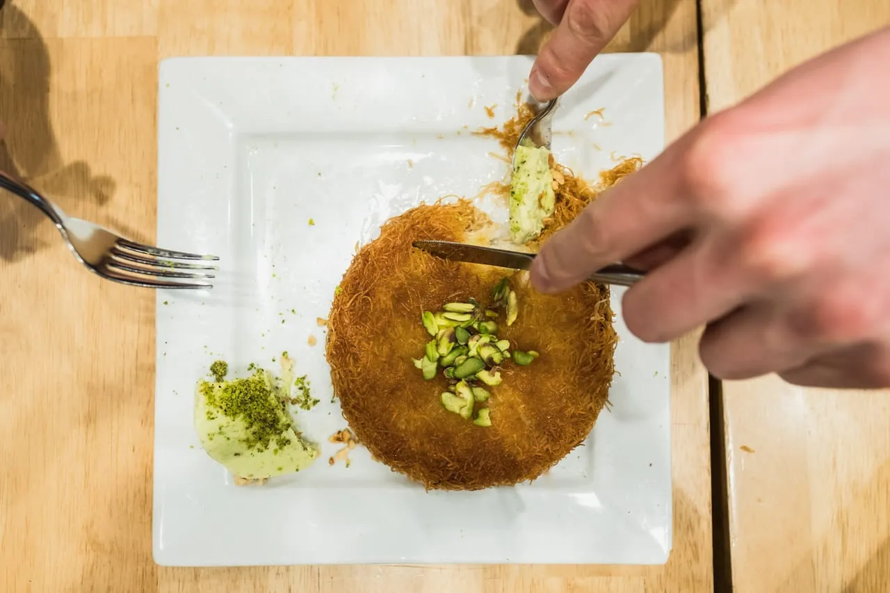 White square plate with crispy kunefe dessert topped with pistachios being cut with knife and fork