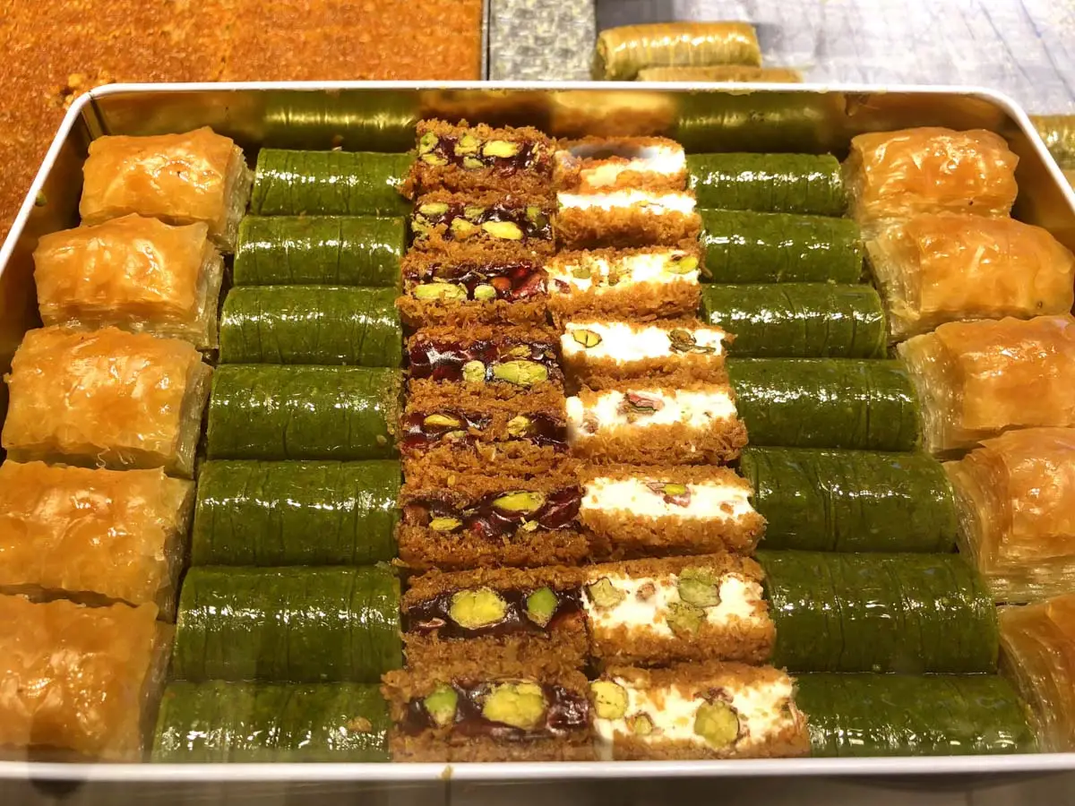 Metal tray packed with rows of green pistachio baklava and golden layered pieces topped with cream
