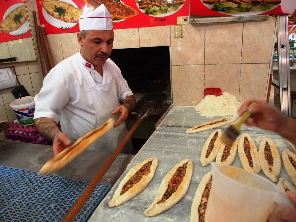 Chef sliding cooked pide from wood-fired oven with large peel while offsider brushes tops with oil