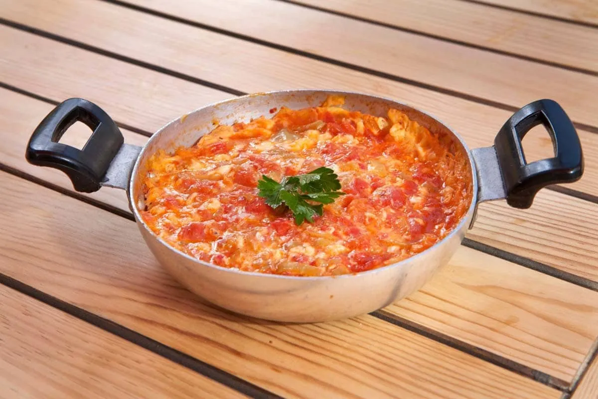 Menemen with scrambled eggs and tomato in a two-handled metal pan on a wooden table