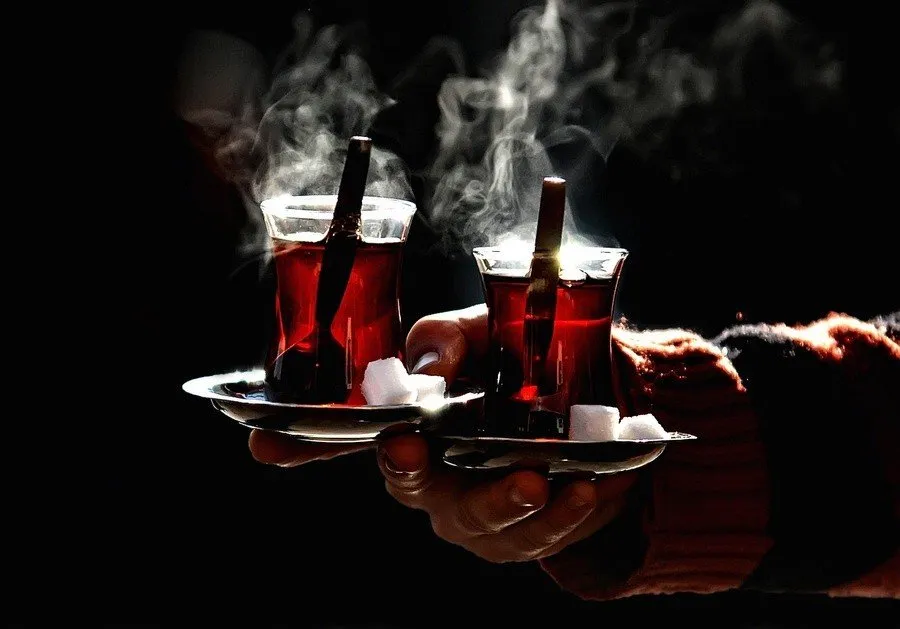 Holding two glasses of red Turkish tea in traditional glasses with spoons and sugar, steam coming off top