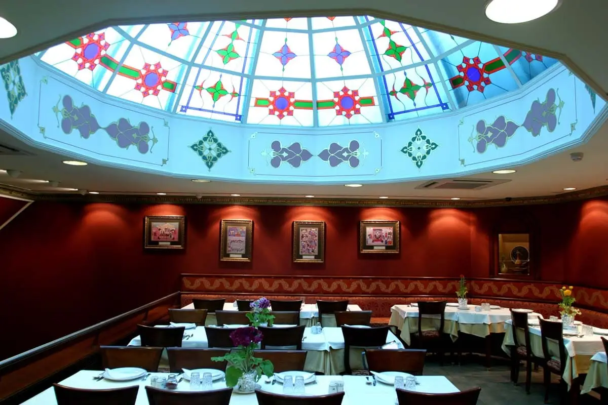 Restaurant interior with a stained glass dome, red walls, framed art, and white-clothed tables set with flowers