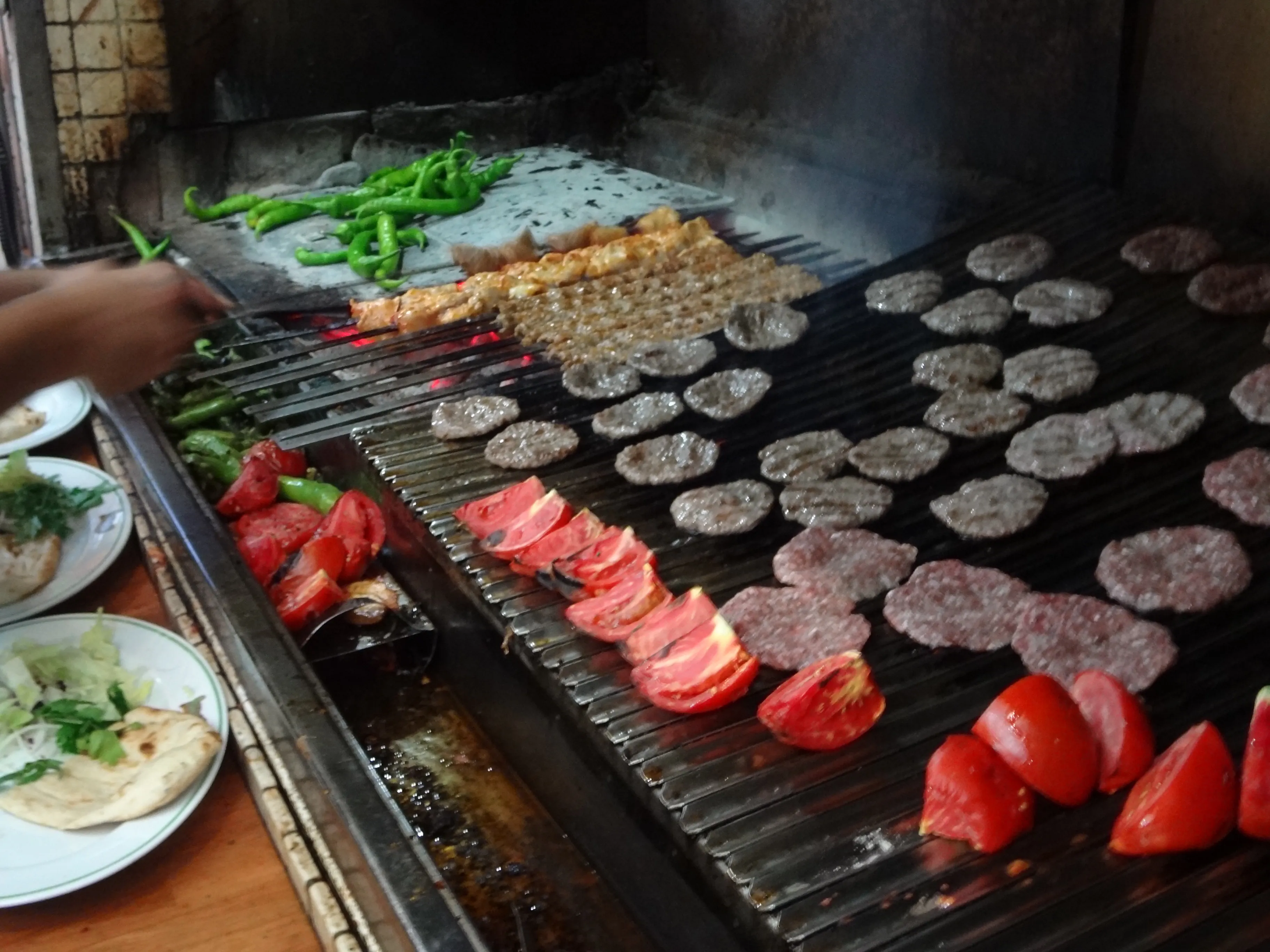 Rows of köfte, skewers, peppers, and tomatoes cooking on a large charcoal grill