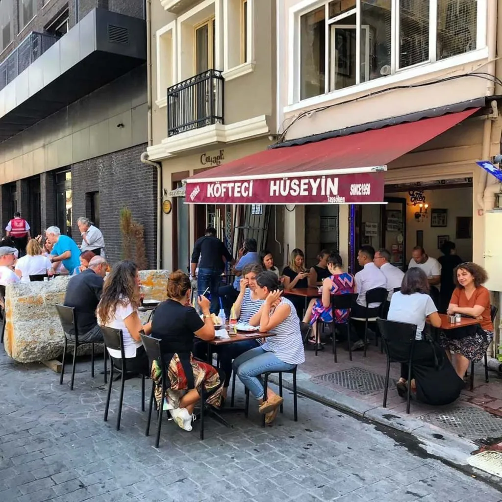 Lunch crowd at sidewalk tables under the red Köfteci Hüseyin awning on Kurabiye Sokak in Beyoğlu