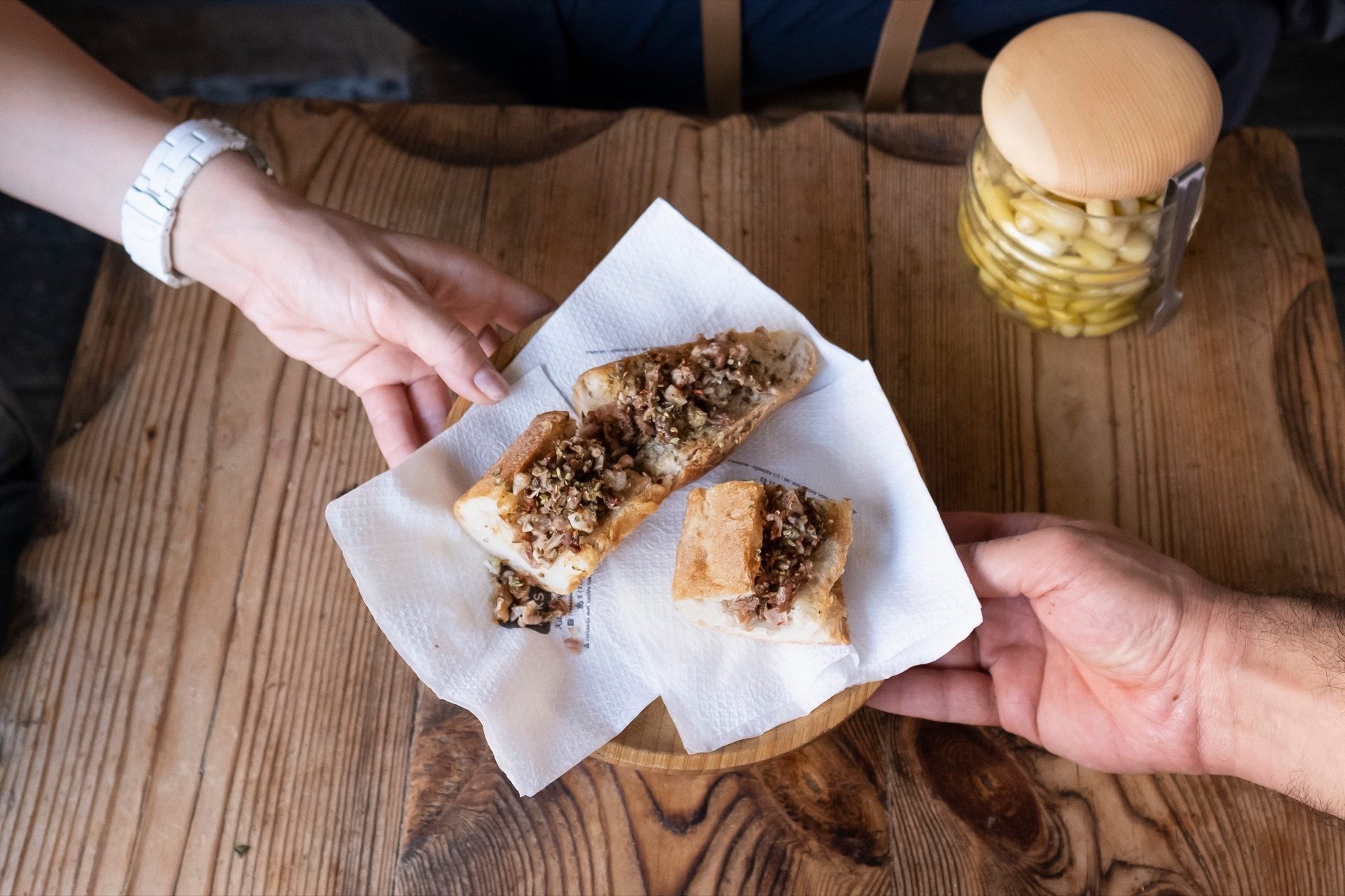 Two hands reaching for kokoreç served on bread, overhead on a wooden board in Istanbul