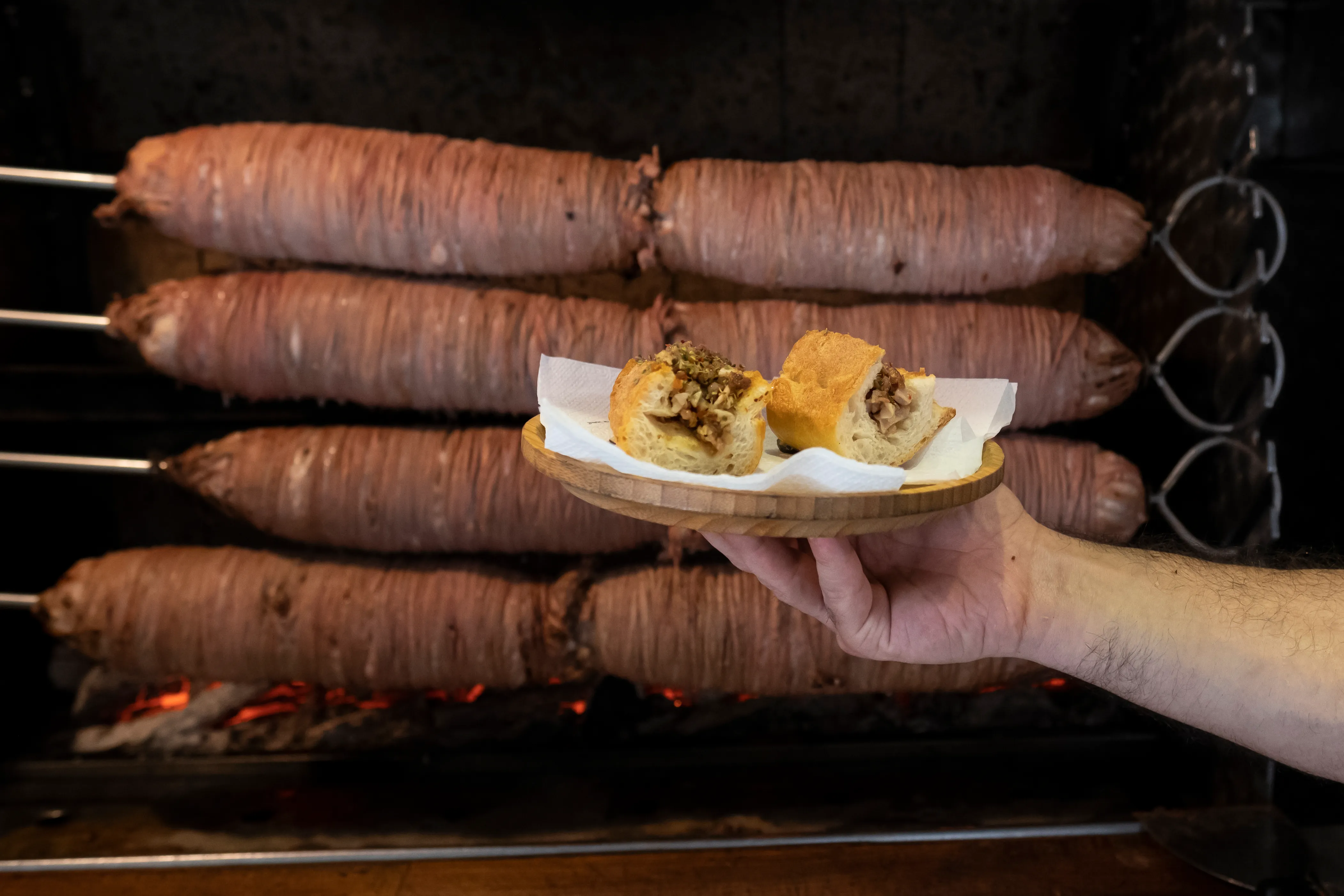 Two kokoreç sandwich halves held on a plate in front of rolls roasting over charcoal