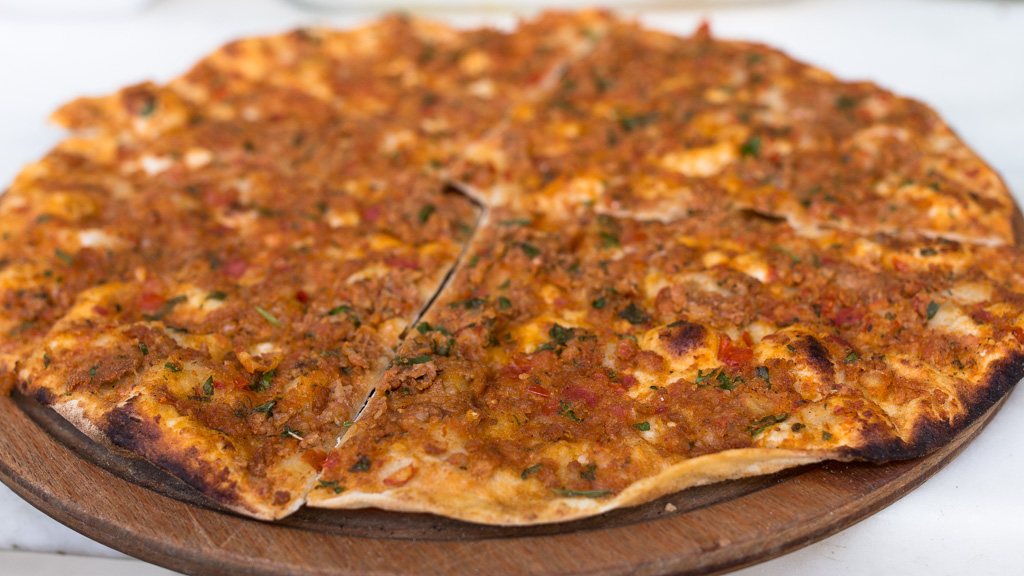 Whole thin-crust lahmacun on a wooden board, sliced into wedges and topped with seasoned minced lamb, fresh parsley and red pepper