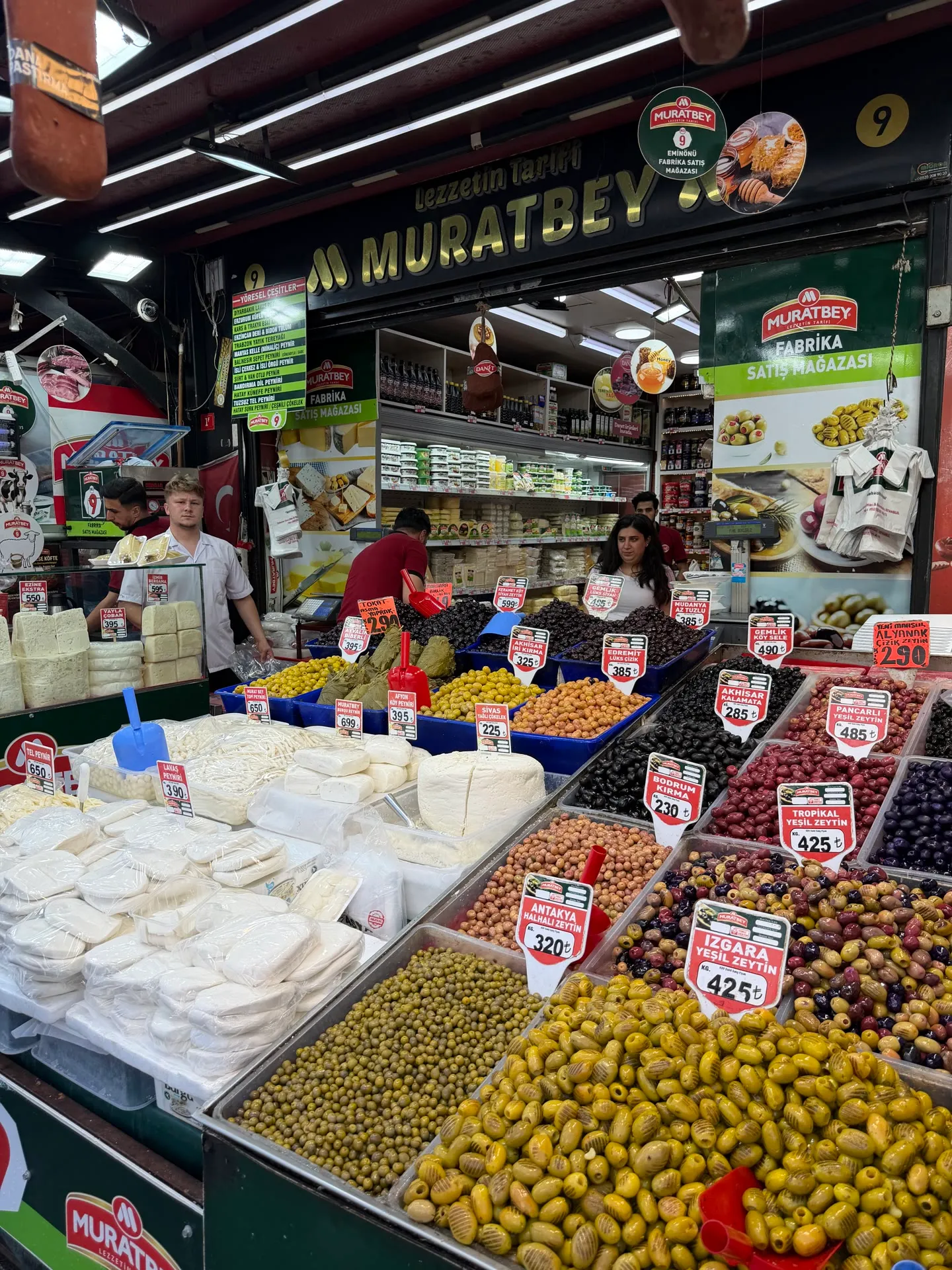 a sarkuteri counter with a dozen varieties of olives in trays and stacked blocks of white cheese