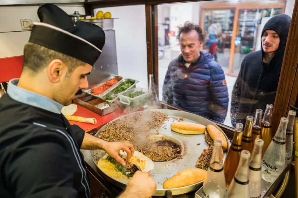 Man in black chef uniform cooking tantuni while two men wait outside window Kadikoy Street Food tour
